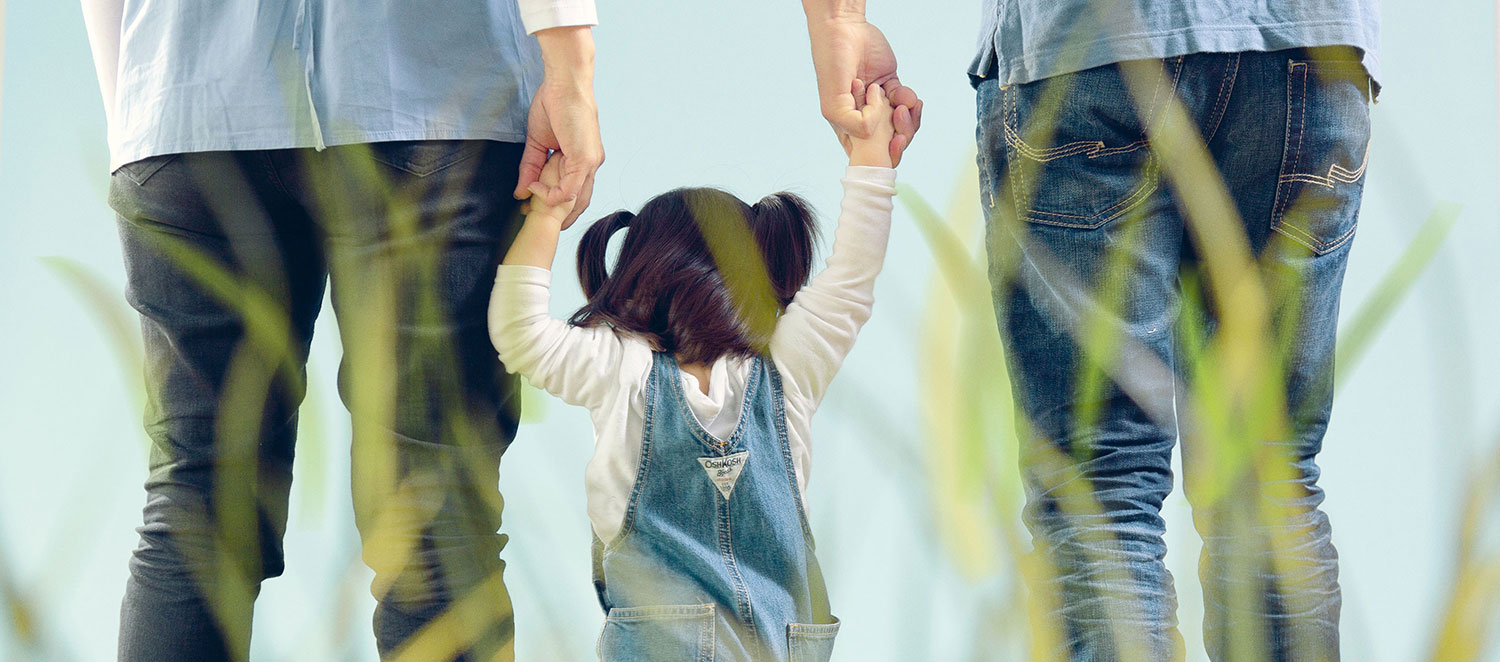 Familie von hinten, zwei Erwachsene halten ein kleines Kind an den Händen