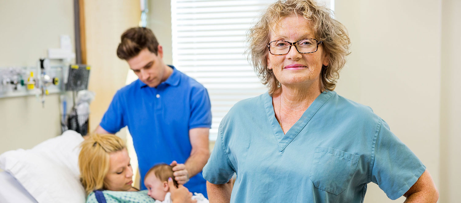 Im Krankenhaus-Zimmer: Lächelnde Hebamme im Vordergrund, im Hintergrund Paar mit Neugeborenem. 