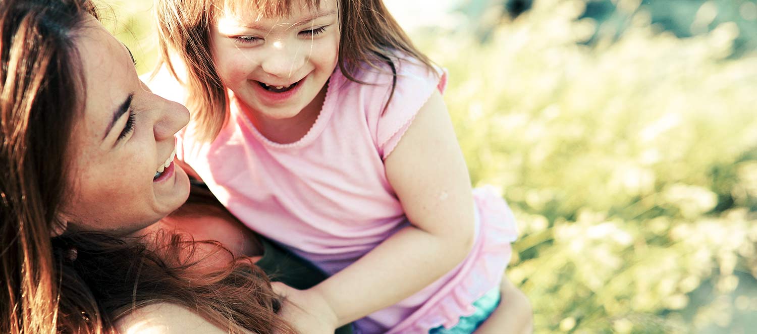 Mutter lachend mit einem Mädchen mit Downsyndrom auf dem Arm. 