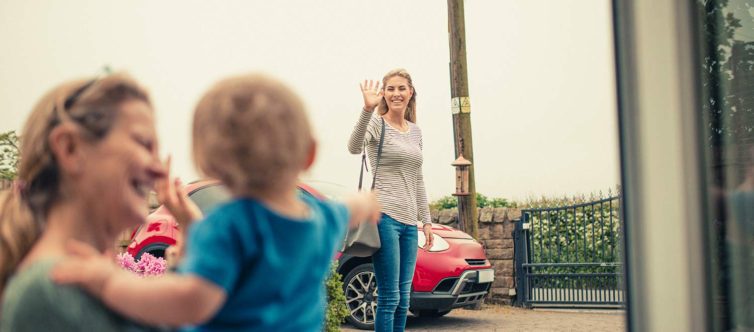 Lächelnde Frau hält Baby auf dem Arm, andere Frau winkt zum Abschied. 