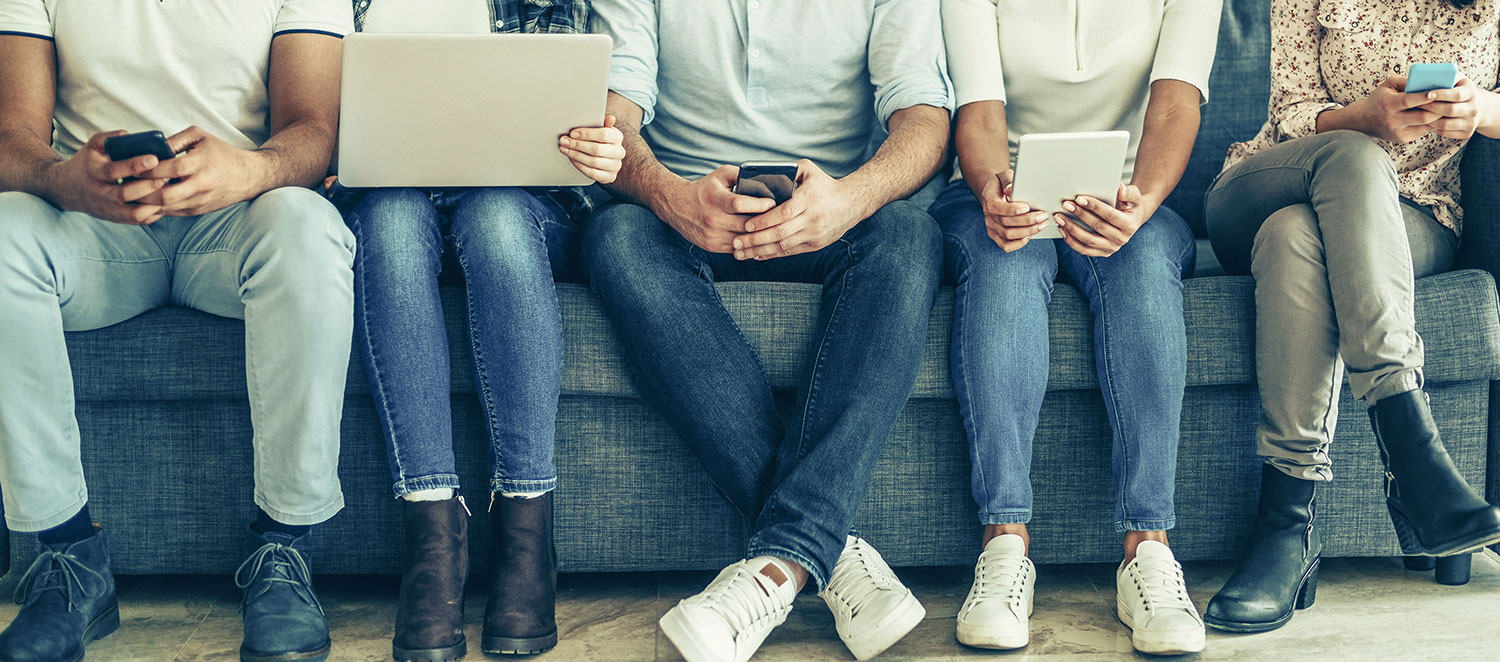 Fünf Erwachsene sitzen nebeneinander auf dem Sofa und bedienen Smartphones, Tablet und Laptop. 