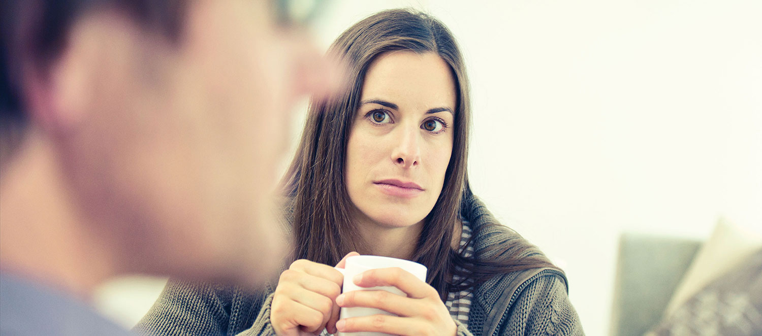 Frau sitzt nachdenklich mit einer Tasse am Tisch, im Vordergrund ist der Partner als Silhouette zu sehen. 