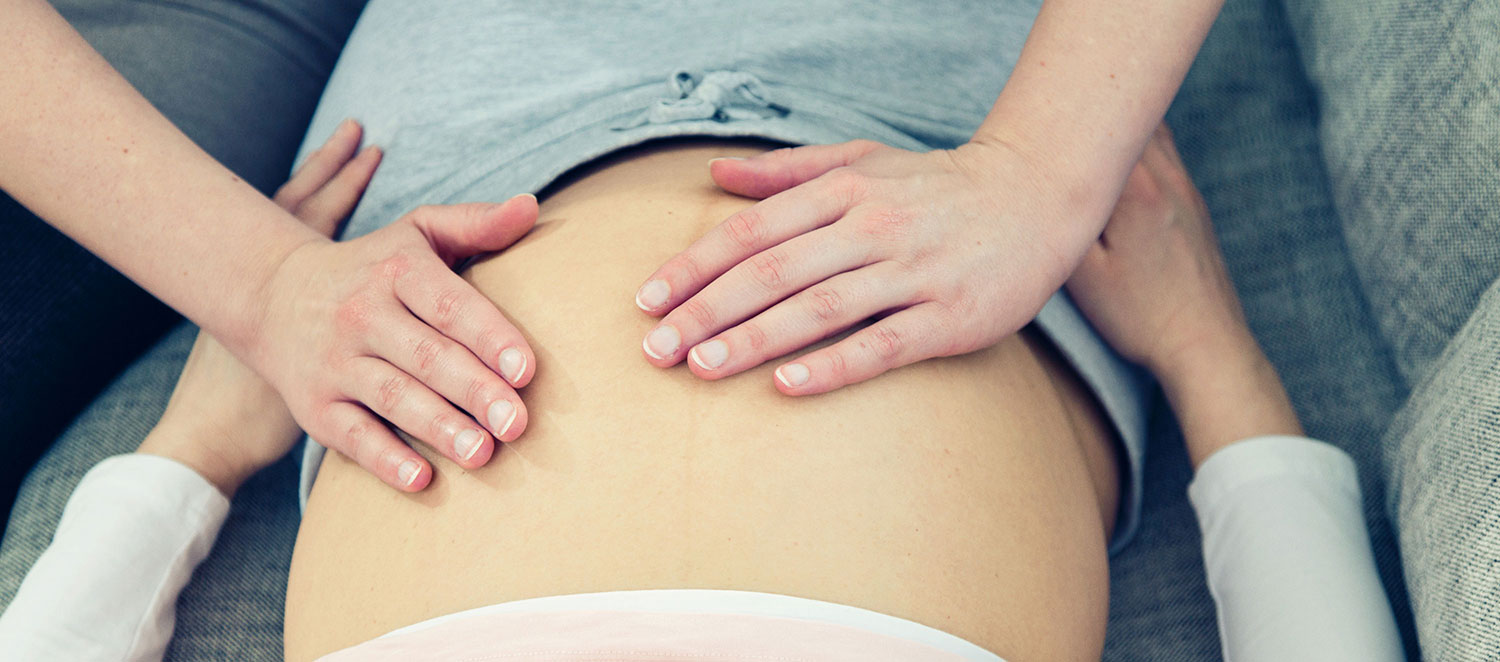 Zwei Hände tasten nackten Bauch einer schwangeren Frau ab. 