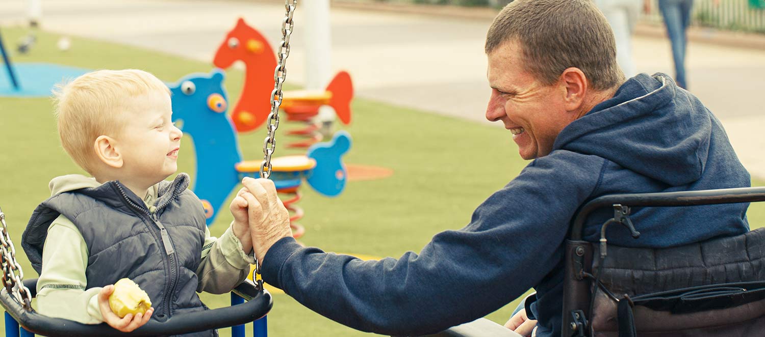 Mann im Rollstuhl spielt mit seinem Sohn auf Spielplatz. 