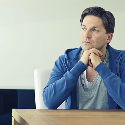 Nachdenklicher Mann sitzt am Tisch, Kopf wird auf verschränkten Händen abgestützt. 