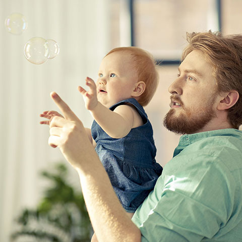 Vater trägt Kleinkind auf dem Arm, beide zeigen auf Seifenblasen. 