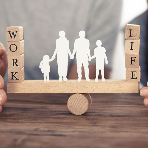 Paar beschützt mit den Händen symbolisch die Work-Life-Balance in Form von Figuren einer Familie auf einer Wippe. 